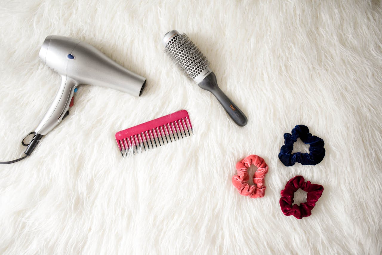 Services-03 Flatlay of hairstyling tools including a hairdryer, brush, comb, and scrunchies on white fur.